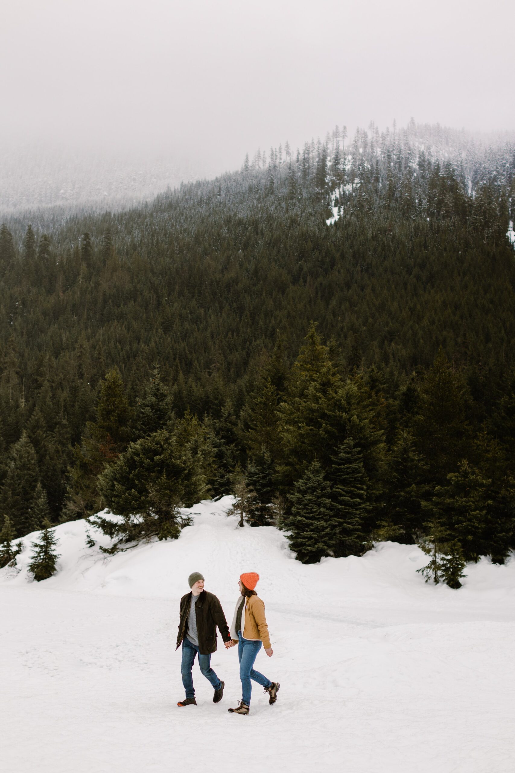 boy and girl walk on snow holding hands with trees rising up behind them during gold creek pond engagement session