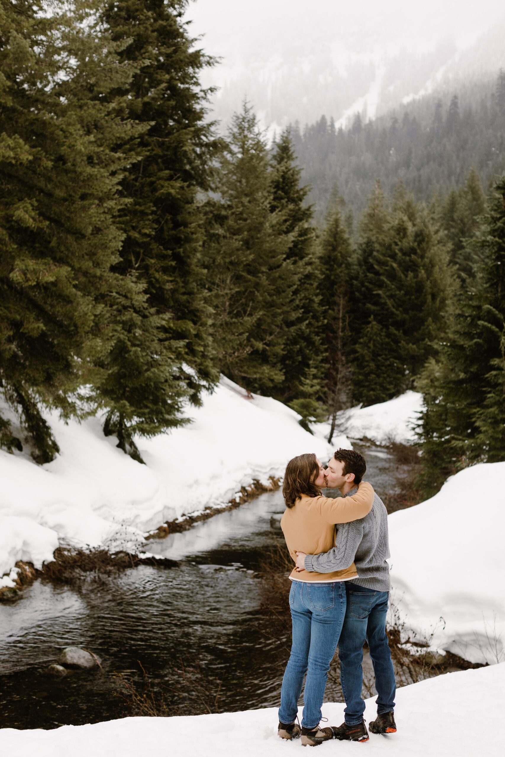 boy and girl embrace in front of river with snow on the ground and evergreens surrounding them