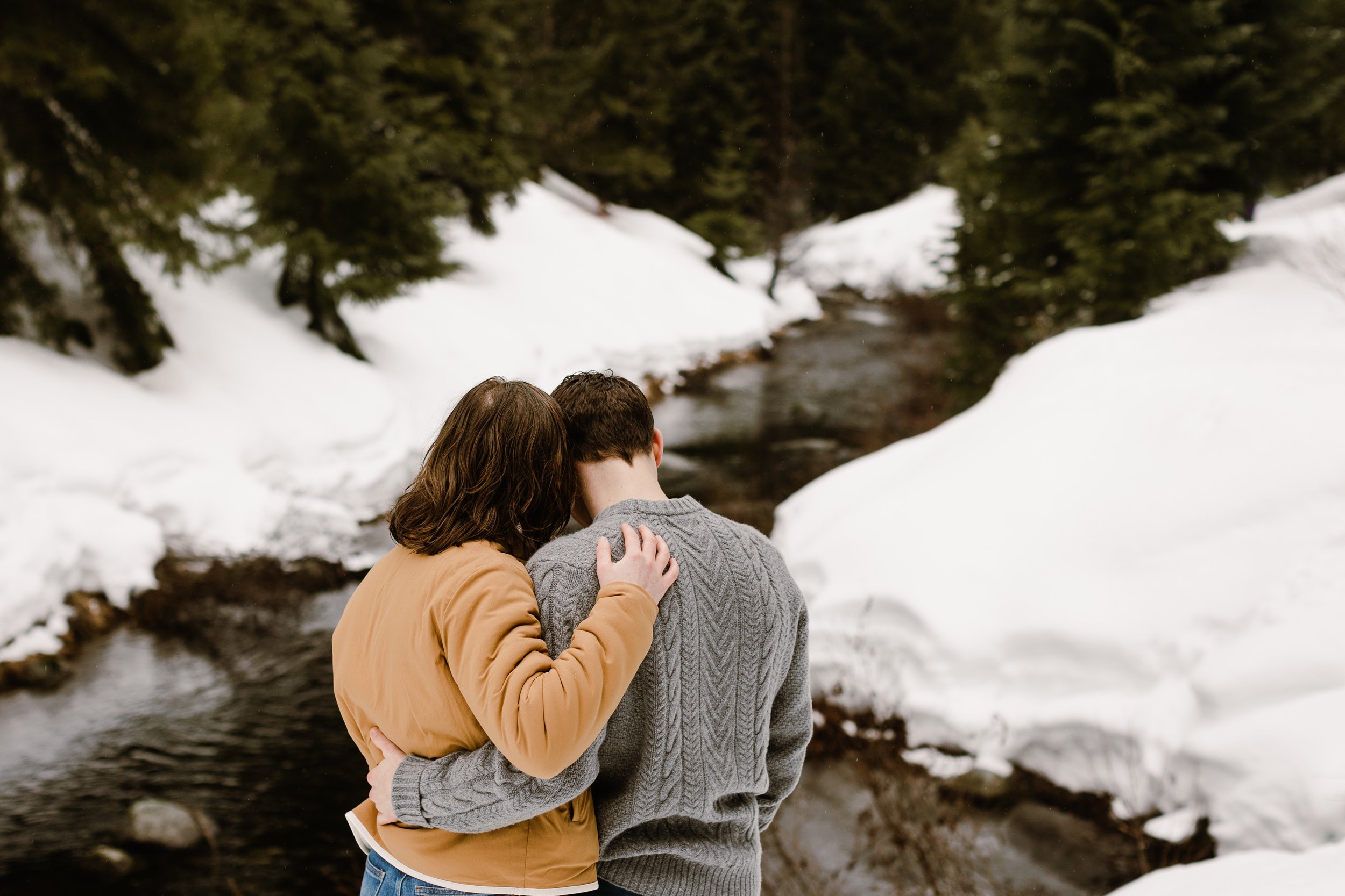boy and girl face a river holding each other with snow and green trees around them