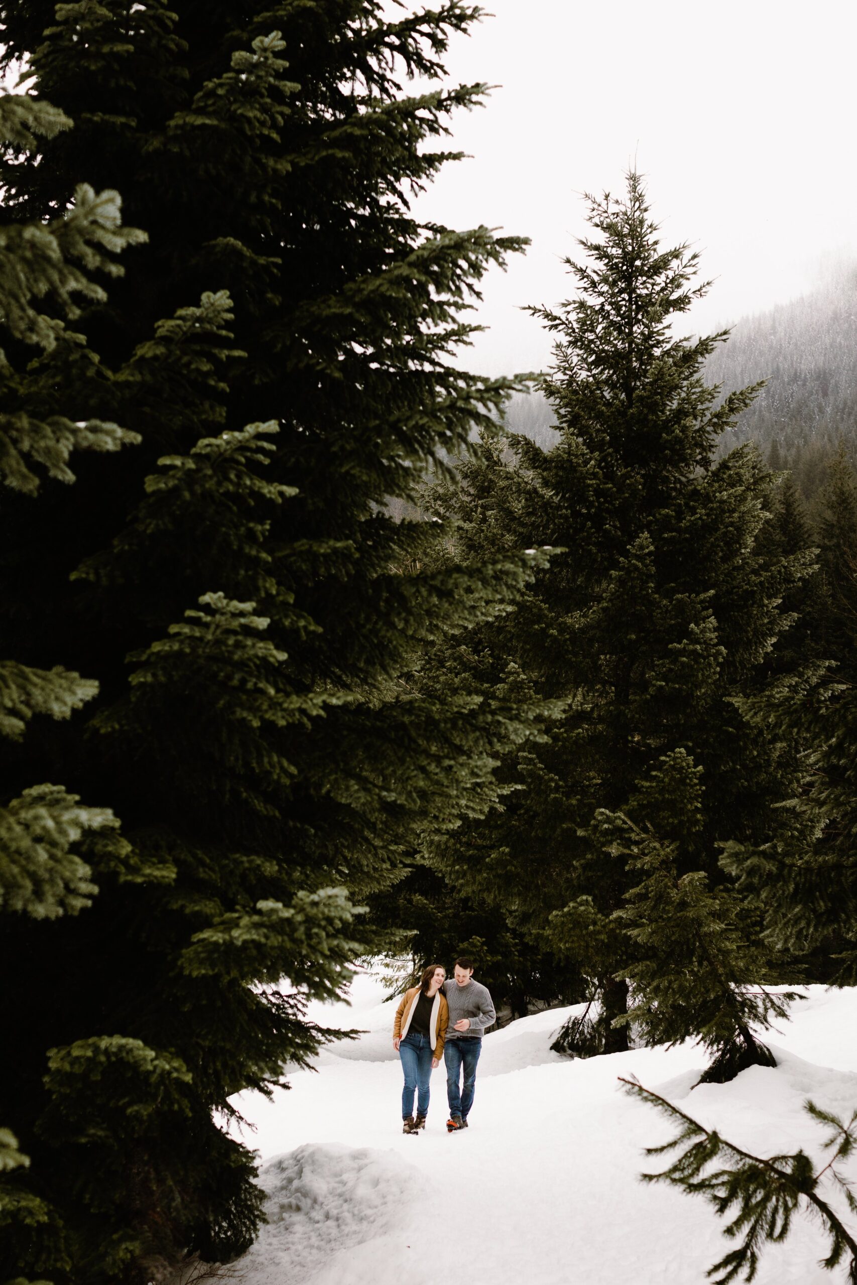 boy and girl walk on snow with trees around them during gold creek pond engagement photo shoot