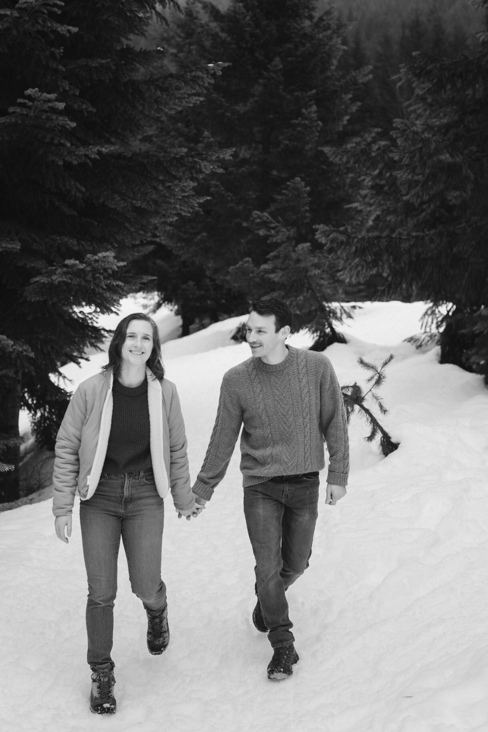 boy and girl walk on snow with trees around them during winter engagement photo shoot