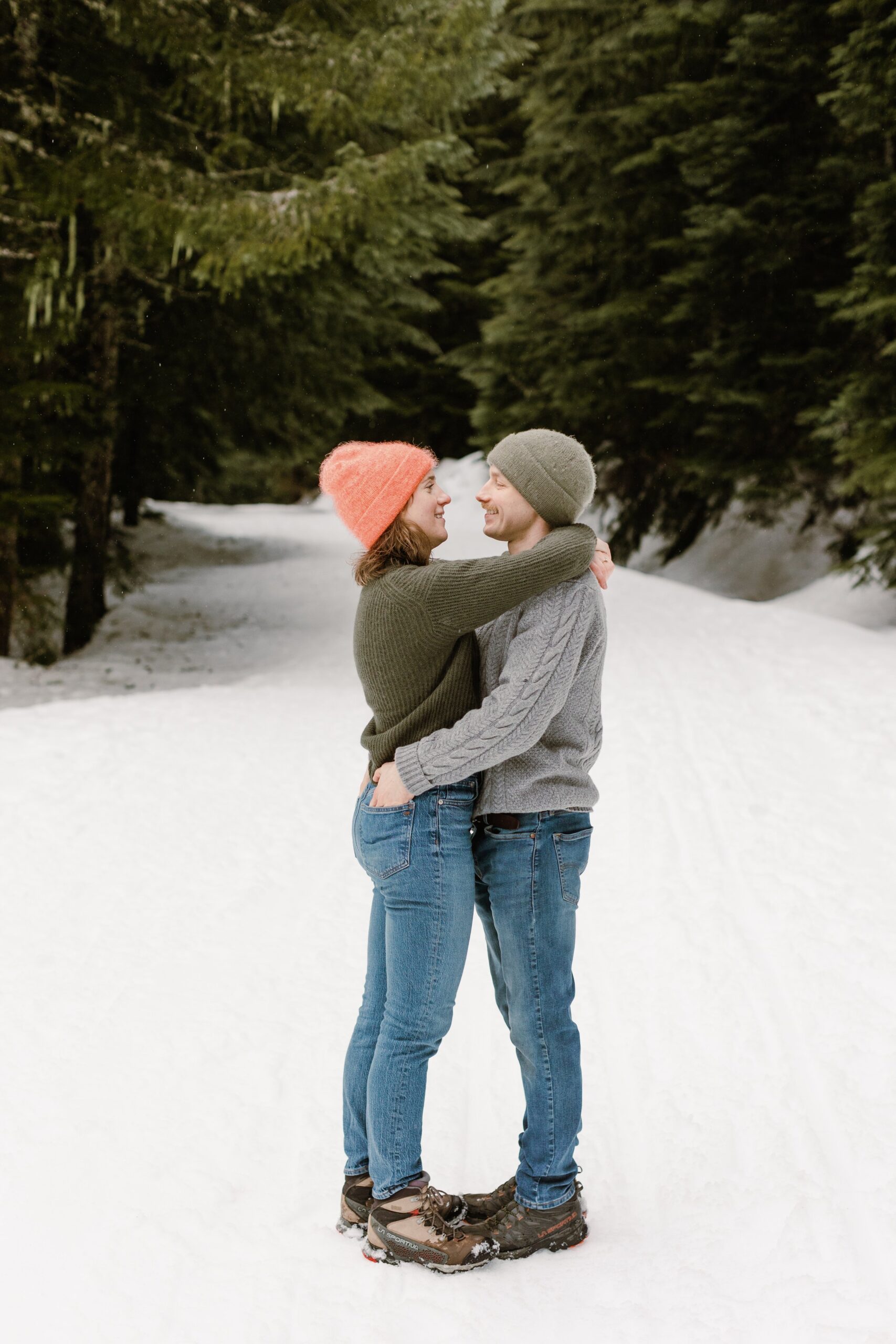 boy and girl embrace while standing on snow and surrounded by evergreen trees