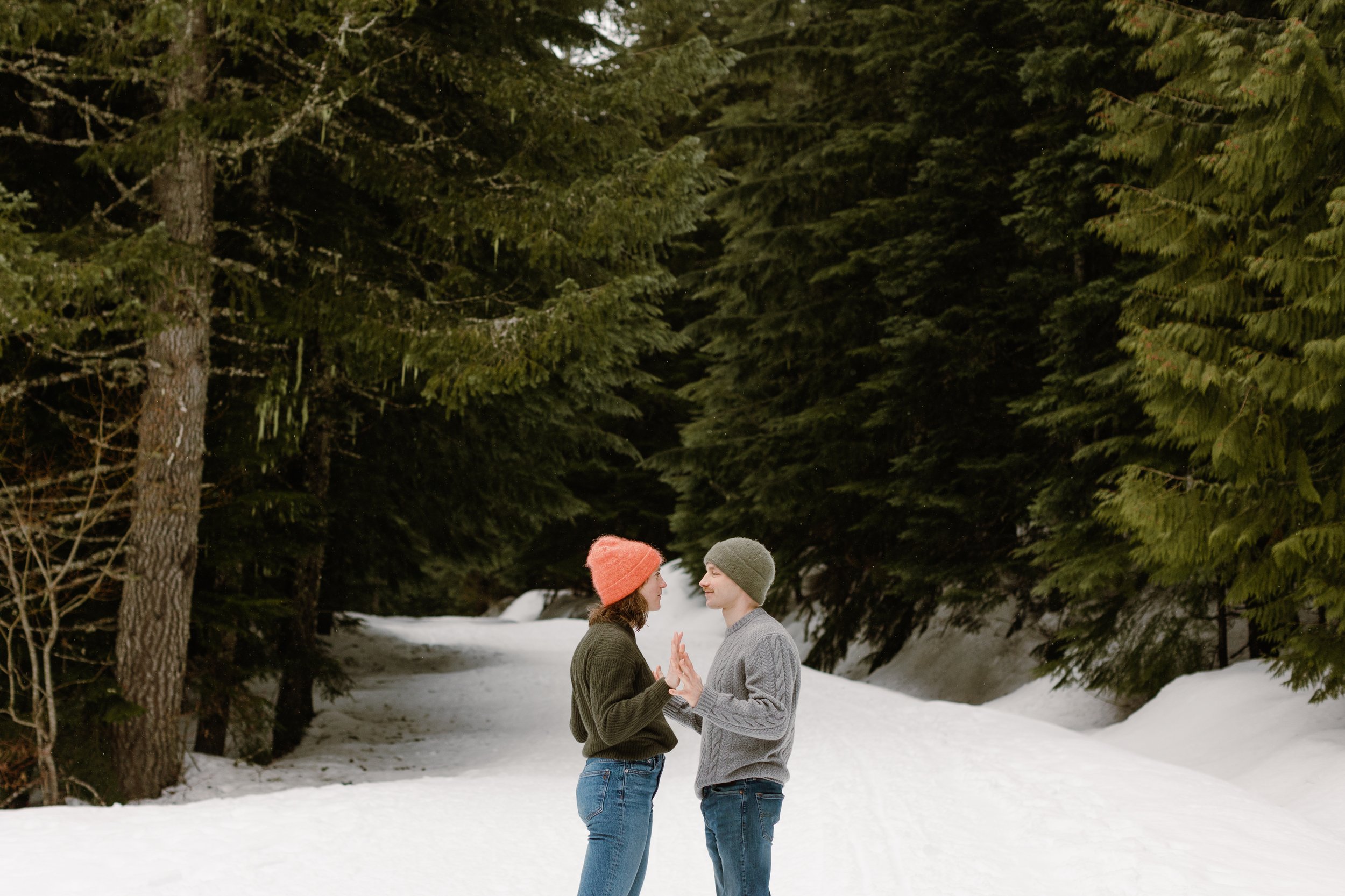 boy and girl face each other with hands touching while standing on snow surrounded by evergreen trees