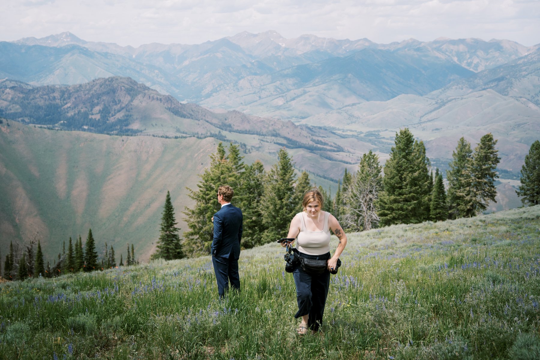 groom standing in a field on a cliffside while photographer walks away from him
